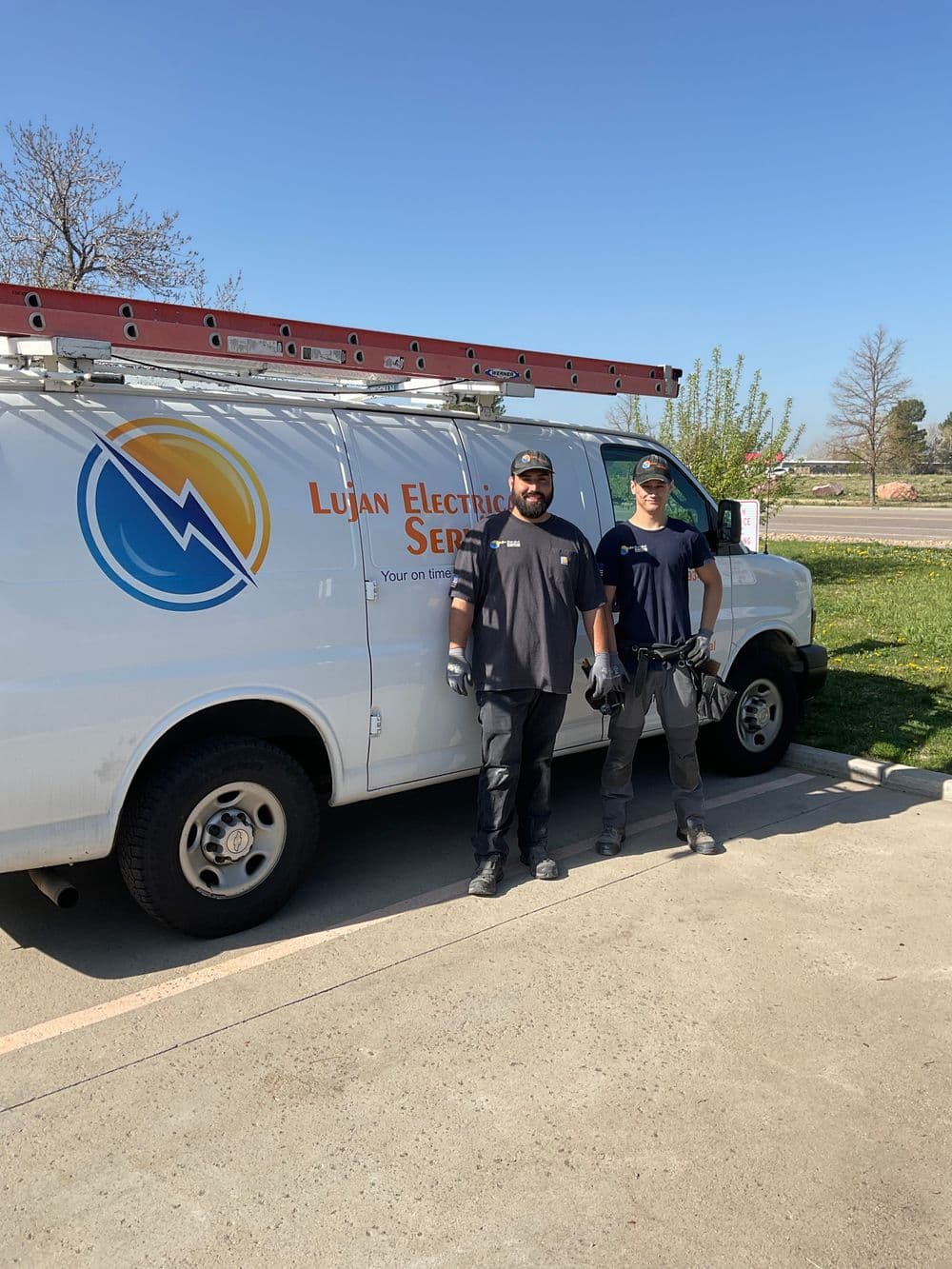 Two electricians standing beside a Lujan Electric Service van on a sunny day.