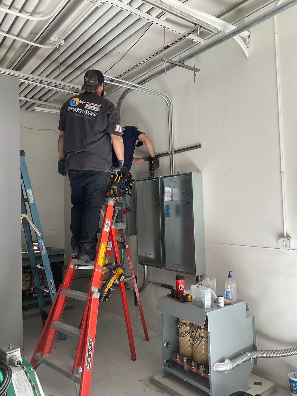 Electricians working on electrical panels using a ladder in a commercial space.