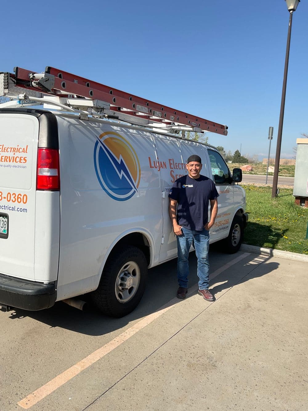 Electrician standing near Lujan Electrical Services van with ladders in a sunny outdoor setting.